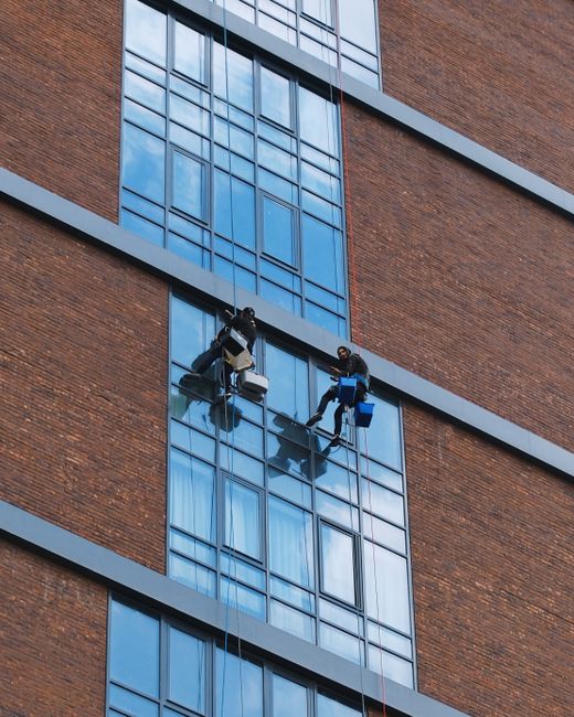 Two professional window cleaners suspended on harnesses working on the exterior glass facade of a tall brick building in Haringey. They are using squeegees and cleaning cloths to maintain the large, reflective glass panels, which are housed within a grid of metal frames. The building features multiple floors with metal horizontal and vertical supports, and natural daylight illuminates the scene. Safety equipment such as harnesses and buckets for cleaning supplies are visible, emphasizing safety and thorough surface cleaning. This image illustrates high-rise window cleaning logistics, highlighting the importance of specialised equipment and professional expertise, as provided by Haringey Carpet Cleaning in their cleaning services context.