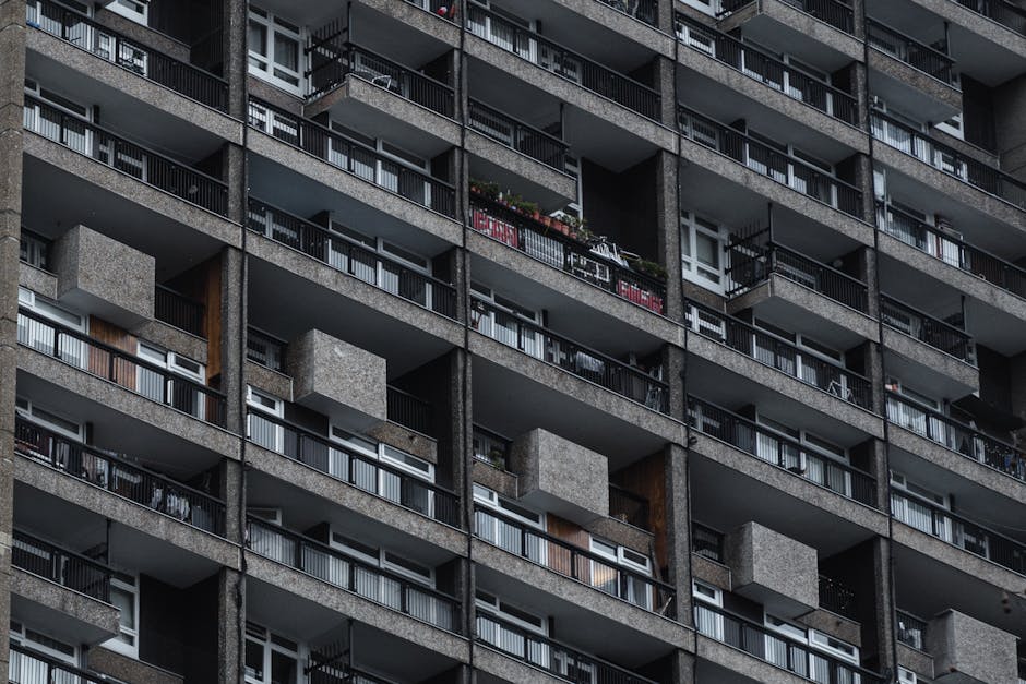 Image of a multi-storey residential high-rise building with numerous balconies, showcasing concrete and metal railings. Several balconies contain potted plants, outdoor furniture, and laundry drying racks. The building's exterior appears weathered, with some faded paint and signs of age. Natural lighting highlights the textured surfaces of the concrete, creating shadows that emphasize the repetitive pattern of the balconies. This exterior shot reflects urban residential architecture, relevant to access challenges and cleaning logistics discussed on the page about high-rise flats in Haringey, with a mention of Haringey Carpet Cleaning for surface and exterior cleaning services.