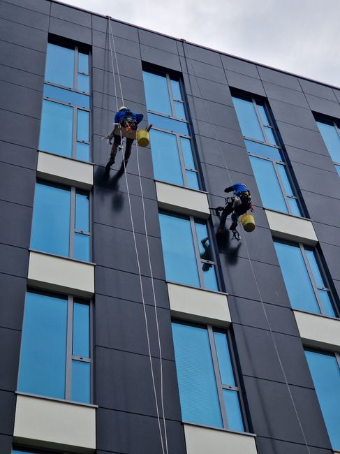 Two professional window cleaners suspended on harnesses working on the exterior glass facade of a tall brick building in Haringey. They are using squeegees and cleaning cloths to maintain the large, reflective glass panels, which are housed within a grid of metal frames. The building features multiple floors with metal horizontal and vertical supports, and natural daylight illuminates the scene. Safety equipment such as harnesses and buckets for cleaning supplies are visible, emphasizing safety and thorough surface cleaning. This image illustrates high-rise window cleaning logistics, highlighting the importance of specialised equipment and professional expertise, as provided by Haringey Carpet Cleaning in their cleaning services context.
