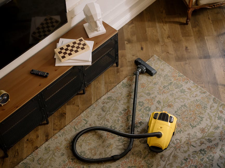 An indoor living room scene featuring a wooden sideboard against a white wall, topped with a white bust sculpture, a chessboard, some papers, a TV remote control, and a small golden clock. The room has hardwood flooring with a patterned, light-colored rug partially covering it. A yellow vacuum cleaner with a black hose and attachment is placed on the rug, ready for surface cleaning or deep cleaning. The room is well-lit with natural light, emphasizing cleanliness and tidy arrangement, consistent with Haringey Carpet Cleaning's professional cleaning services.