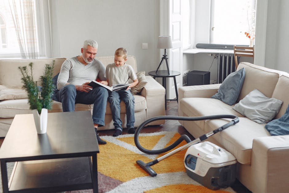 A living room with white walls and large windows letting in natural light. There are two beige sofas, one with a blue cushion and the other with a white cushion. A small black side table with a lamp and a potted plant is positioned between the sofas. On the carpeted floor, a vacuum cleaner with a black hose is visible, indicating ongoing surface cleaning. The room features a wooden floor under the carpet, which shows a clean, well-maintained appearance. The space appears tidy and well-organized, reflecting professional domestic cleaning standards. Haringey Carpet Cleaning's logo or branding is not visible in the image, but the setting emphasizes cleanliness and maintenance, aligning with the focus on avoid common mistakes when booking carpet cleaning services.