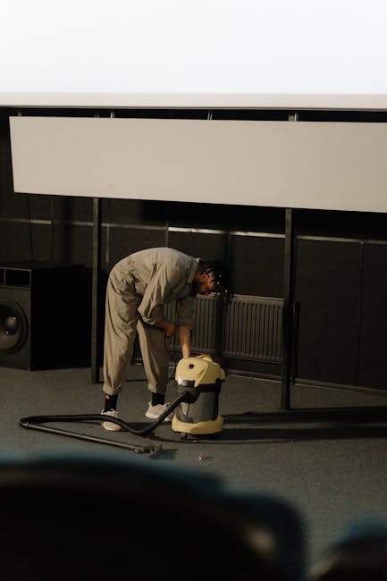 A person performing surface cleaning with a vacuum cleaner on a dark carpeted floor in a professional setting, possibly a conference room or auditorium. The individual is bent over, focused on vacuuming, and is dressed in light-colored work attire. Behind them, there is a black wall with a grey radiator and a large white projection screen or panel above. The room features subdued lighting, highlighting the cleanliness and maintenance work being carried out, consistent with professional cleaning services offered by Haringey Carpet Cleaning. The scene emphasizes thorough carpet cleaning as part of deep cleaning and hygiene maintenance.