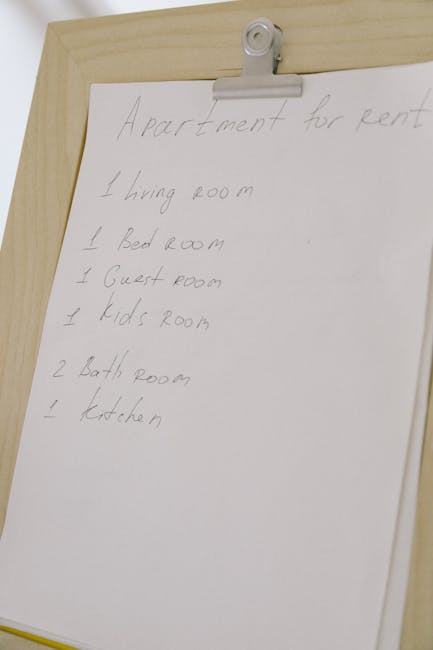 A white laminated sheet with handwritten details of an apartment layout, including rooms such as the living room, bedroom, guest room, kids' room, bathrooms, and kitchen, clipped onto a wooden surface with a metal clip at the top. The background shows a slightly textured wooden surface. This image appears to relate to preparation for end of tenancy cleaning and surface organisation, highlighting room listings for cleaning services provided by Haringey Carpet Cleaning. The scene is well-lit with natural lighting, emphasizing the simplicity and clarity of the handwritten notes.