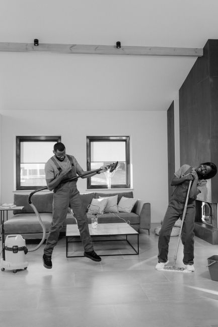 Two professional cleaning staff members from Haringey Carpet Cleaning stand back-to-back against a plain white wall, dressed in beige uniforms. One woman holds a vacuum cleaner with a red hose and black body, while the other is holding a green-handled mop, both smiling confidently. The background is plain, emphasizing their neat, well-maintained cleaning tools. The scene underscores the importance of thorough surface cleaning and hygiene, aligning with the theme of avoiding hidden charges in cleaning quotes by showcasing professionalism and comprehensive domestic cleaning services provided by Haringey Carpet Cleaning.