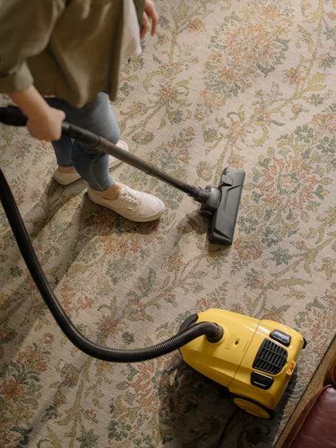 A child in beige shoes and jeans is using a vacuum cleaner with a grey and yellow body to clean a patterned rug in a living room. The rug has a floral design in muted tones of green, beige, and orange. The vacuum cleaner's hose is visible, connecting to the handheld nozzle. The room is well-lit with natural or artificial light, and the setting appears tidy and well-maintained. Haringey Carpet Cleaning offers professional surface cleaning and deep cleaning services to ensure optimal hygiene and cleanliness for domestic spaces.