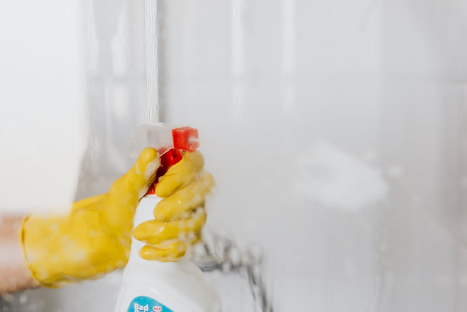 Close-up of a person wearing yellow rubber gloves using a white spray bottle to clean a vertical tiled glass surface, likely a shower or bathroom mirror, in a well-lit room. The tiles are glossy and light-colored, with some water spray visible on the surface. The background shows a clean, modern bathroom or kitchen setting with bright natural light. The image emphasizes surface cleaning and sanitisation, aligning with Haringey Carpet Cleaning's same-day cleaning solutions offered in Haringey, UK.