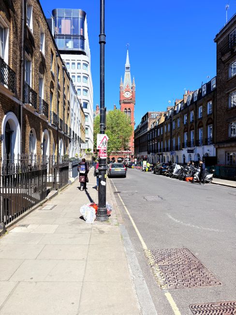 A city street scene with a paved sidewalk and a row of brick and stone residential buildings on the right, featuring large windows and balconies. On the left, there is a fence with a row of small trees and a woman walking on the sidewalk, carrying a bag. Street parking includes several parked motorcycles and cars, and a street sign is visible. The sky is bright blue with a clear view of a historic red brick clock tower with a tall spire in the background, surrounded by modern high-rise buildings. The image depicts an urban environment in daylight, with no cleaning activity visible. Haringey Carpet Cleaning's website, haringeycarpetcleaning.co.uk, emphasizes domestic and commercial cleaning solutions, but this specific street scene does not show cleaning processes.