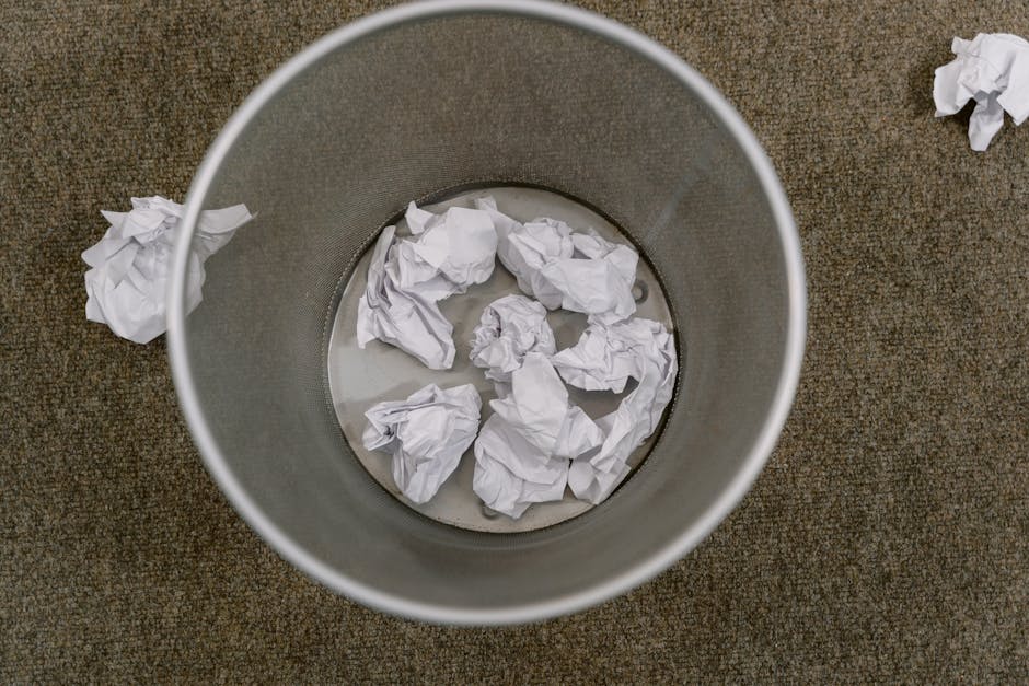 Top-down view of a round, metallic rubbish bin with a smooth surface, containing several crumpled white paper balls submerged in water. Surrounding the bin are additional crumpled white papers scattered on a textured brown carpeted floor. The lighting is natural and even, highlighting the cleanliness of the surfaces, with no visible dirt or debris, indicating a focus on waste management and surface cleaning as part of domestic or commercial cleansing routines. The setting appears to be an office or home environment, with the bin positioned centrally in the image associated with waste disposal, emphasizing hygienic disposal practices. Haringey Carpet Cleaning is mentioned naturally within the context of maintaining hygienic standards associated with cleaning services.