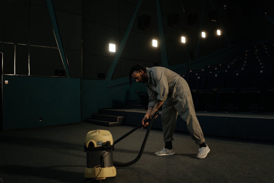A person wearing a light grey jumpsuit and white sneakers is performing deep cleaning with a yellow and black vacuum cleaner on an empty, darkened auditorium floor. The floor appears to be a dark carpet, and the room features tiered seating with numbered rows, illuminated by overhead lighting. The walls are blue with angled panels, and a small staircase leads to the higher rows. The environment suggests professional cleaning of a large public or event space, emphasizing surface cleaning and hygiene maintained by [COMPANY_NAME] for optimal sanitisation and upkeep, consistent with guidelines for waste removal and cleanliness on the page 
