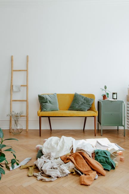 A bright, minimalist living room featuring a yellow fabric upholstered sofa with two green cushions, positioned against a plain white wall. To the left, a wooden ladder-style shelving unit holds a few decorative items and a small plant. On the right, a light green side table with a vintage design supports a potted plant, a picture frame, and a small electronic device. The wooden parquet floor is scattered with crumpled and unfolded clothes in various colors, indicating recent domestic cleaning or tidying activity. Natural lighting from an unseen window illuminates the space, emphasizing the clean, tidy aesthetic. This scene represents a typical setting where professional cleaning specialists, like those from Haringey Carpet Cleaning, may perform surface cleaning and deep cleaning services to maintain hygiene and freshness in residential spaces, especially in the context of sofa and room cleaning near Wood Green N22.