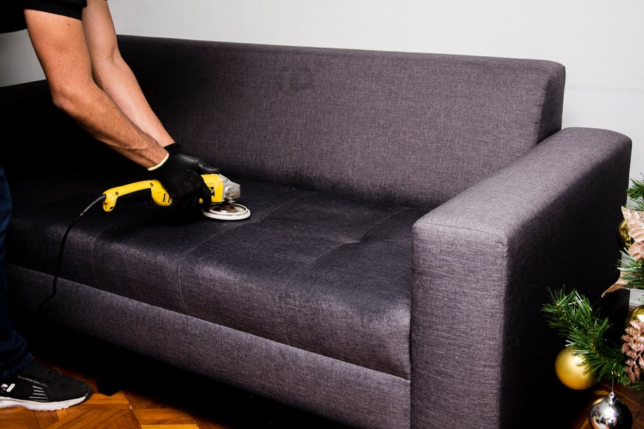 A person wearing black gloves is performing surface cleaning on a dark grey fabric sofa in a residential living room, using a handheld electric upholstery cleaner to deep clean the sofa cushions. The sofa is positioned against a white wall, with natural light illuminating the space, and a Christmas decoration with green foliage and gold baubles partially visible on the right side of the image. The scene showcases careful hygienic maintenance of domestic furnishings, emphasizing professional sofa sanitisation and deep cleaning services provided by Haringey Carpet Cleaning, a trusted provider near Wood Green N22.